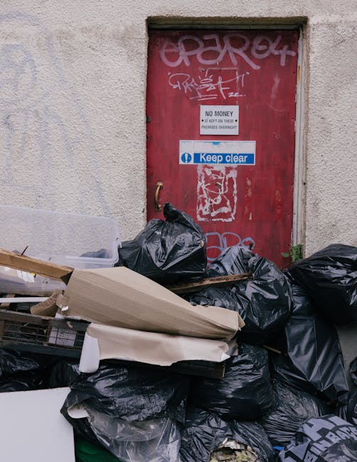 A pile of black rubbish bags, some with visible creases and shiny textures, stacked in front of a red metal door that is covered in graffiti tags and stickers. The door features a white sign stating 'NO MONEY' and a smaller notice indicating 'Keep clear,' with faded paint and graffiti overlapping these signs. Adjacent to the bags, an assortment of discarded items including cardboard boxes, a piece of foam, and a wooden plank with nails partially sticking out is visible, suggesting an informal waste collection or clearance scene. The scene is set against a rough, off-white exterior wall, with some small patches of greenery at the base of the wall, indicating an urban environment. The overall atmosphere is observational, highlighting a typical instance of rubbish that might be associated with private waste removal services such as those offered by Rubbish Clearance Chelsea, illustrating an alternative approach to waste management outside of local authority collection. Natural lighting casts minimal shadows, emphasizing the textures and details of the refuse and the door's surface.
