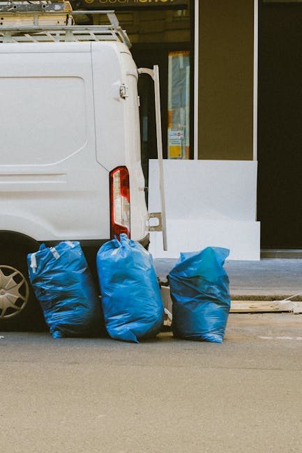 The image shows the rear section of a white commercial van parked on a city street, with three large blue rubbish bags placed on the ground immediately beside it. The bags are made of thick plastic material, slightly crinkled, and filled with waste, with some surface folds and creases visible. The van's tailgate and rear lights are partially visible, with the rear door closed. Behind the van, there is a building with a glass window or door, and a white rectangular panel leaning against the building's entrance. The scene suggests a private waste collection or rubbish removal activity, possibly arranged through an independent rubbish clearance service such as Rubbish Clearance Chelsea, serving customers seeking alternative disposal options away from local authority bins. The overall lighting indicates daytime, with natural light illuminating the scene, and the setting appears to be an urban area with a clean, paved street surface. The arrangement of rubbish bags and the presence of the van imply a scheduled or on-site waste removal process.