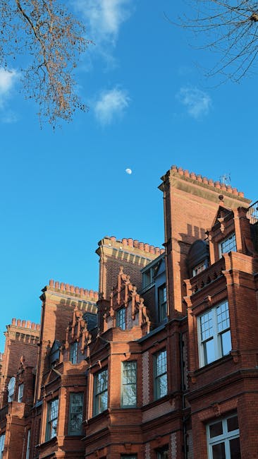 The image depicts a clear blue sky with a visible crescent moon high above a row of historic red-brick residential buildings, characteristic of Victorian architecture, with ornate gables, decorative battlements, and large sash windows. The buildings occupy the lower portion of the frame, showcasing their detailed brickwork and stone accents, while the upper sections feature protruding bay windows and intricate crenellations along the rooftops. In the top left corner, tree branches with sparse, dry foliage extend into the shot, contrasting against the bright sky. The scene appears to be in a quiet urban setting, possibly in Chelsea, with natural daylight illuminating the textured brick surfaces. The scene is neutral and professionally composed, reflecting a calm, residential environment. Although the image does not explicitly show rubbish or waste, it presents an architectural context where professional rubbish removal services, such as those offered by Rubbish Clearance Chelsea, might support on-site clearance or private disposal needs for such properties.