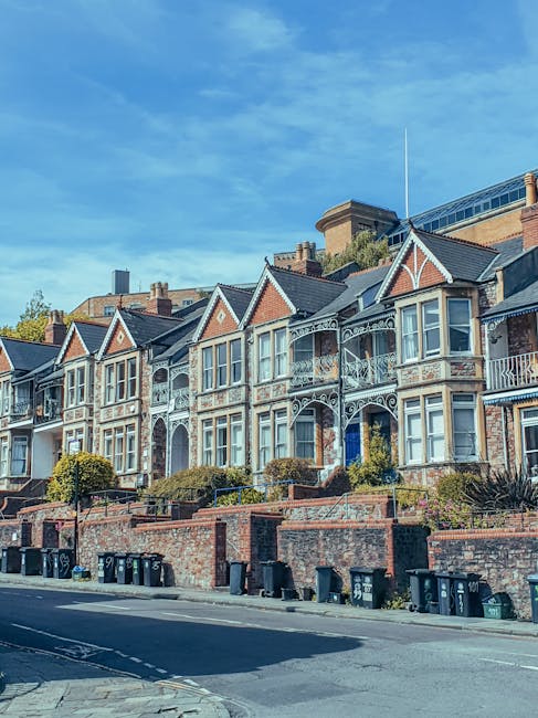 A row of Victorian-style terraced houses situated on a slightly elevated street. The houses feature ornamented ironwork balconies, gabled roofs with decorative trim, and large bay windows. The façades are constructed from brick with some painted details, and the front gardens are bordered by low brick walls with steps leading up to the entrances. In front of the houses, there is a paved pavement and a street lined with multiple black wheelie bins, which are likely used for rubbish or recycling collection. The background includes modern buildings and a clear blue sky with a few scattered clouds, suggesting a sunny day. The scene presents a typical residential street scene, where private waste and rubbish management could be involved, highlighting the importance of professional rubbish clearance services in maintaining tidy urban environments.