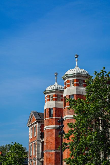 The image depicts a clear blue sky with a visible crescent moon high above a row of historic red-brick residential buildings, characteristic of Victorian architecture, with ornate gables, decorative battlements, and large sash windows. The buildings occupy the lower portion of the frame, showcasing their detailed brickwork and stone accents, while the upper sections feature protruding bay windows and intricate crenellations along the rooftops. In the top left corner, tree branches with sparse, dry foliage extend into the shot, contrasting against the bright sky. The scene appears to be in a quiet urban setting, possibly in Chelsea, with natural daylight illuminating the textured brick surfaces. The scene is neutral and professionally composed, reflecting a calm, residential environment. Although the image does not explicitly show rubbish or waste, it presents an architectural context where professional rubbish removal services, such as those offered by Rubbish Clearance Chelsea, might support on-site clearance or private disposal needs for such properties.