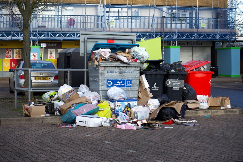 A large collection of mixed waste, including cardboard boxes, black and red trash bags, paper and plastic packaging, and discarded household items, piled on the pavement outside a commercial building. The waste is partially spilling onto the cobblestone street, with some items leaning against a metal railing. Behind the heap, there is a blue recycling bin labeled for mixed paper and cardboard, positioned next to a smaller black bin, both overflowing with rubbish. To the left, a silver car is parked next to the overflowing waste, with trees and a retail storefront with signage visible in the background. The scene suggests an instance of non-collection or improper rubbish disposal, in an urban area that may be serviced by private waste removal providers such as Rubbish Clearance Chelsea, supporting alternative waste management options outside council collection schedules. The environment appears outdoor and well-lit, offering a clear view of the accumulation of waste in a commercial or retail district.
