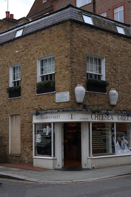 A three-story brick building situated at the corner of a street, featuring white-framed sash windows on the upper floors, each adorned with dark green window boxes filled with small plants. The facade has a weathered brown brick finish, with a sloped black slate roof on the top level. On the ground floor, there is a white storefront with large glass windows displaying interior signage for a business named Chelsea Green, as well as a white entrance door slightly offset to the right. Above the entrance, a white awning extends outward, bearing the business name in black lettering, with additional signage visible inside through the windows. Two large ceramic pots, painted white with a matte finish, hang symmetrically on the brick wall just above the storefront. To the left of the building, a small white street sign indicates the corner of Gough Street SW3 and Grafton Street SW3. The setting appears to be a quiet urban area with a paved sidewalk and a sloped road in the foreground. The scene is captured in natural daylight, highlighting the textures of the brickwork and the details of the storefront, with subtle shadows cast by the building elements. This image relates to private property and commercial premises, which may require independent rubbish removal or on-site clearance services from companies like Rubbish Clearance Chelsea, supporting alternative waste handling that is common in urban environments.